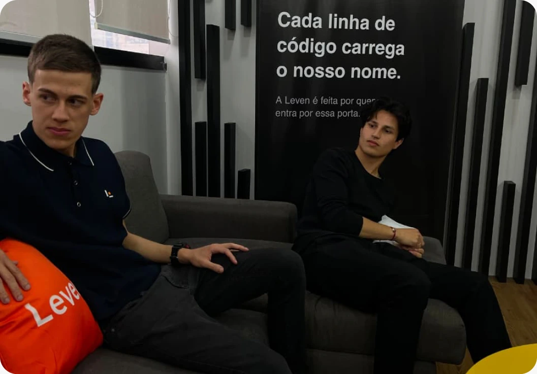 Duas pessoas sentadas no sofá do escritório da Leven, diante do painel "Cada linha de código carrega o nosso nome"