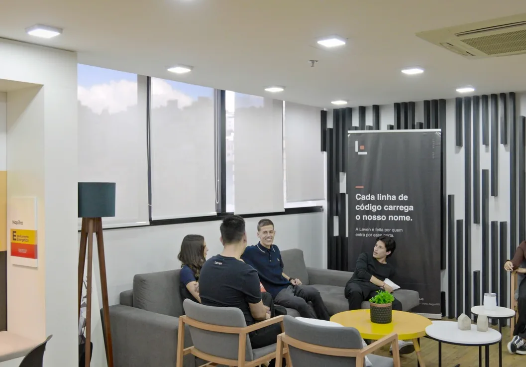 Área de convivência do escritório da Leven, com o time conversando nos sofás e o painel da marca ao fundo